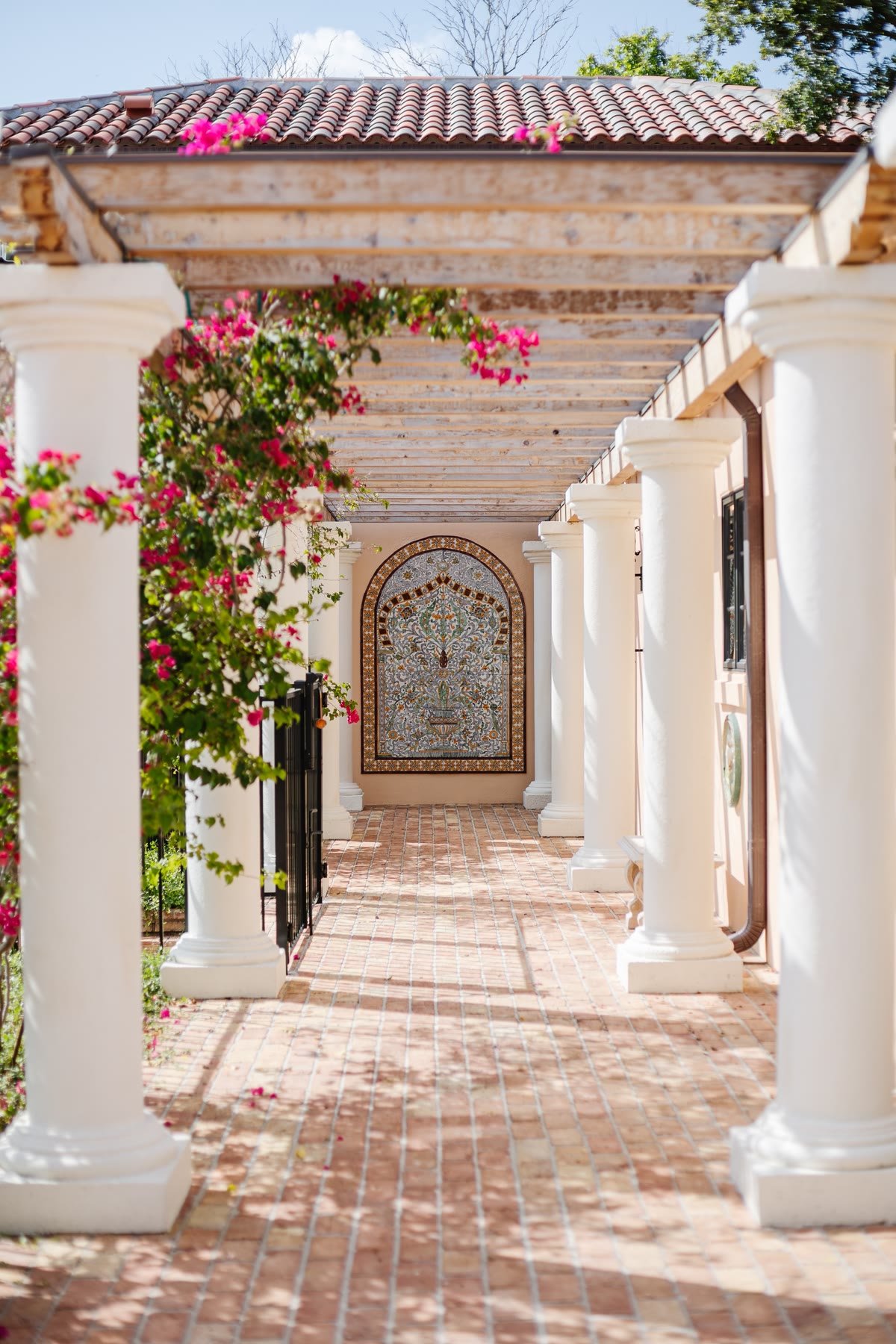 White-columned colonnade with a tiled mural, wooden pergola, and bougainvillea