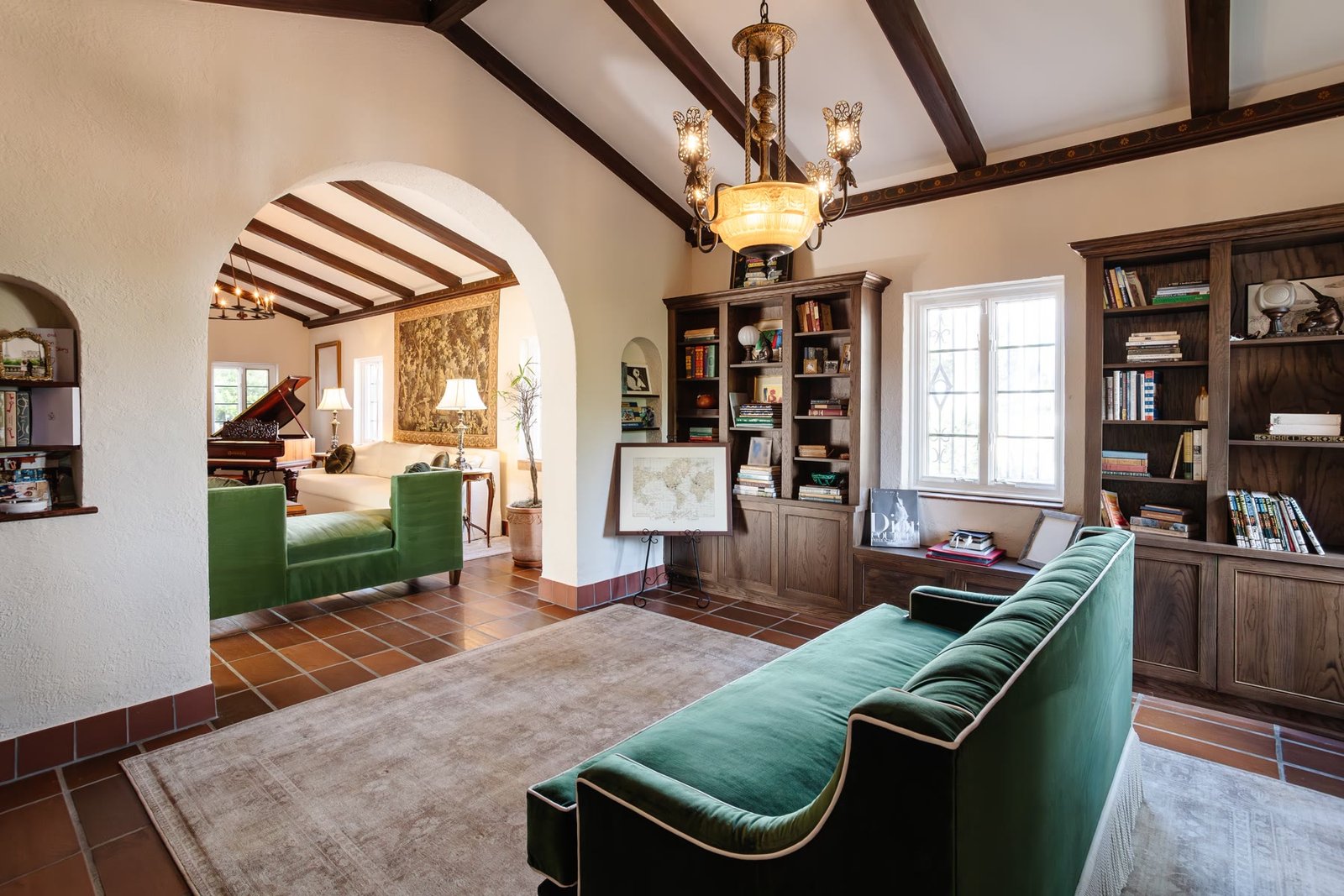Restored library with terracotta floors, dark beamed ceiling, built-in millwork, a green velvet sofa, and an arched opening to the piano room